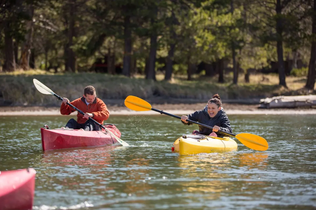 Two kayakers paddle across a serene body of water surrounded by lush, green trees. The scene captures the thrill of outdoor adventure and the beauty of Central Oregon's natural landscape.