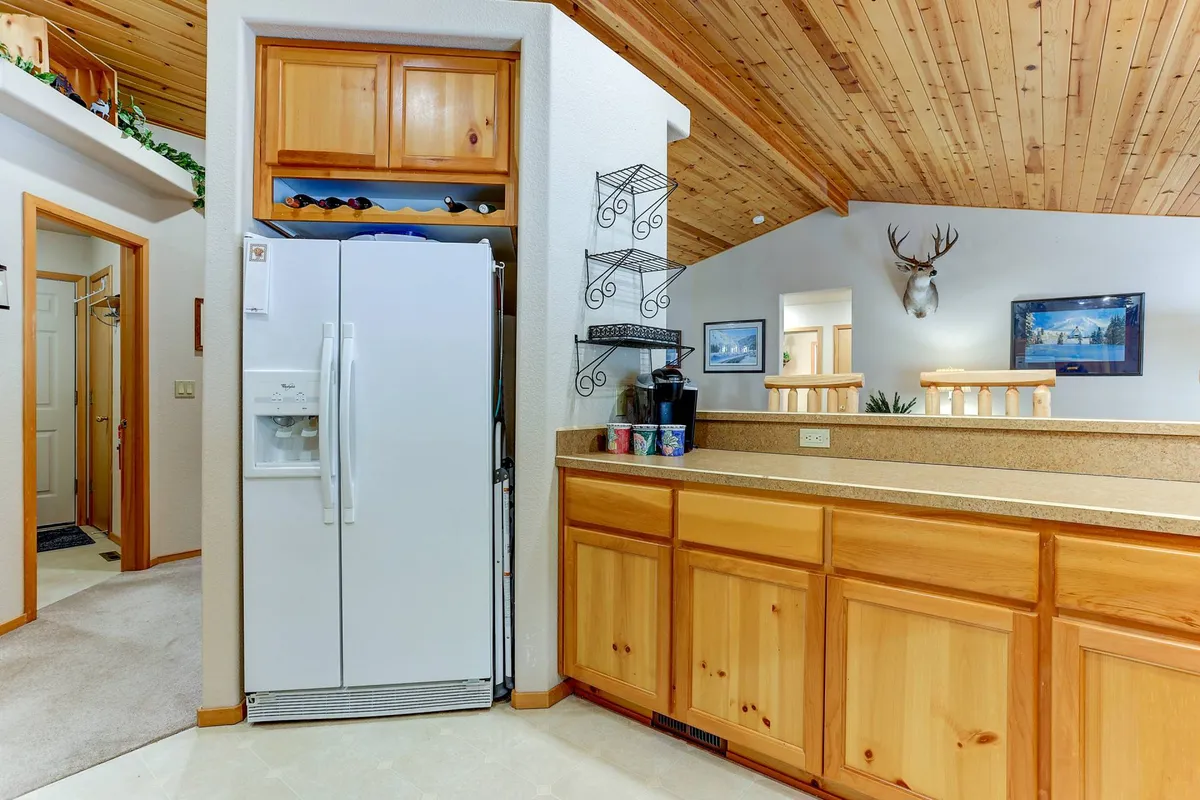 A welcoming kitchen scene featuring a refrigerator, spacious counter, wooden cabinets, and a decorative rack with cups. Antler decor adds rustic charm, and the layout encourages both meal prep and casual gatherings. The view hints at ample room for both cooking and conversing.
