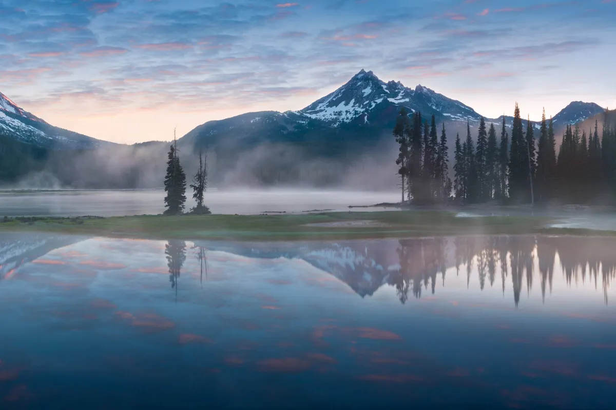 A panoramic view of mountains, a tranquil lake, and encircling trees under a cloud-speckled sky. The landscape suggests opportunities for hiking, exploring, and nature appreciation.