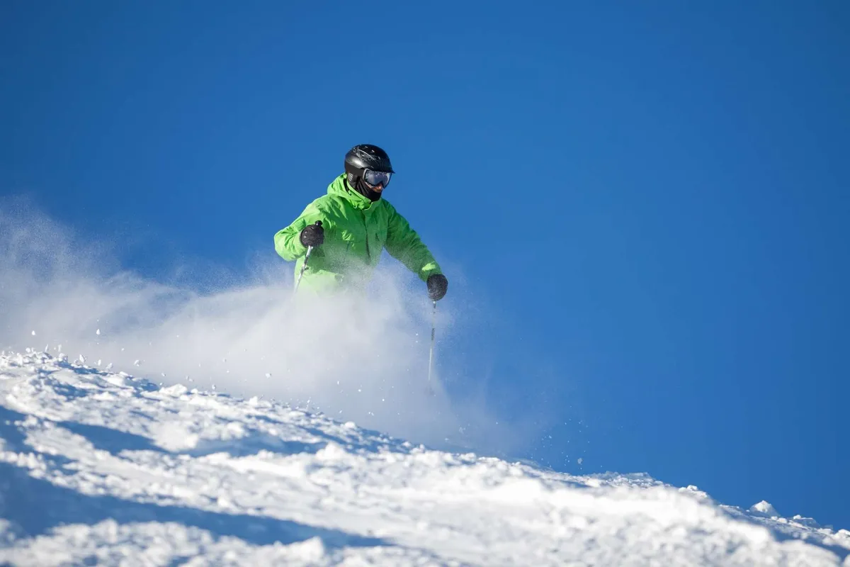 A skier in a bright green jacket glides down a snow-covered slope beneath a clear blue sky, capturing the thrill and winter beauty of the surrounding landscape. The scene emphasizes the proximity to world-class winter recreation at Mt. Bachelor, with an expansive view of snow-draped pines and mountain terrain.