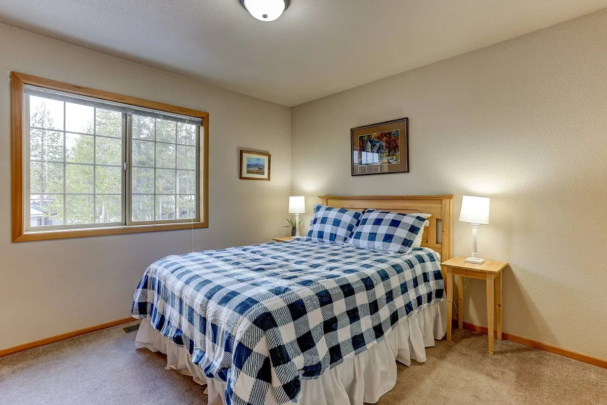 This image shows a serene bedroom with a bed featuring plaid bedding, a window offering natural light and a pleasant view, a nightstand with a lamp, and framed artwork on the wall. The inviting ambiance is ideal for relaxation and restful sleep.