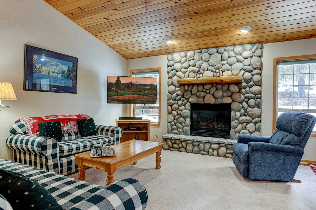 This image showcases a cozy living room with a striking stone fireplace as the centerpiece, flanked by inviting plaid sofas and a warm wooden coffee table. The space exudes a lodge-inspired charm, perfect for gathering with loved ones, relaxing by the fire, or enjoying a movie night after a day of adventure in Sunriver.