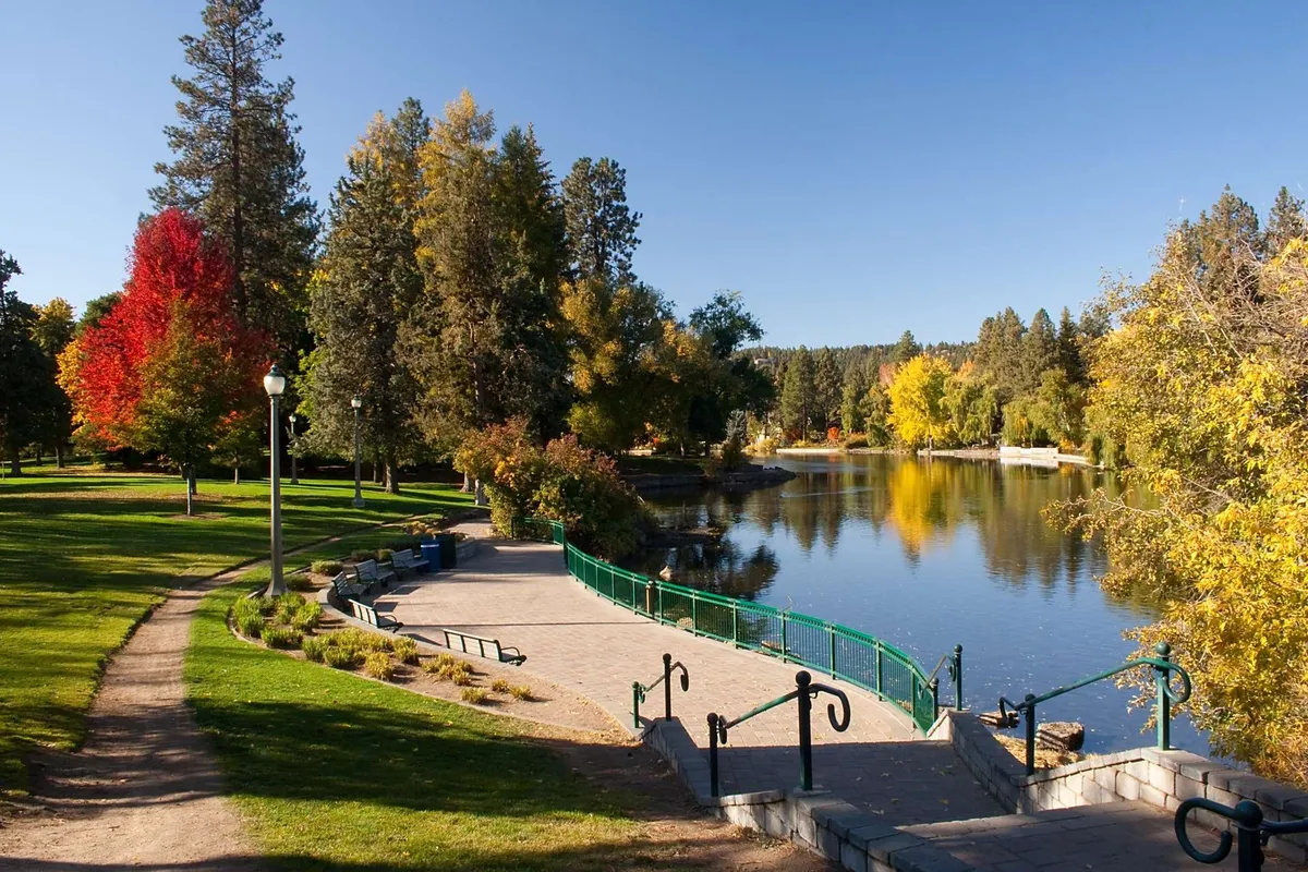 A serene river winds through a vibrant park, with walking paths, colorful trees, and a green railing. The landscape invites strolling, picnicking, and casual exploration.