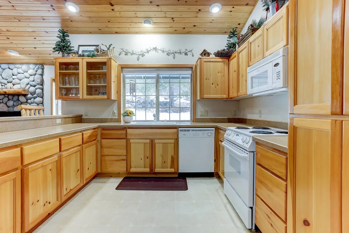 This image showcases a cozy kitchen with wooden cabinetry, a large window allowing natural light, and a stone wall feature. The scene is filled with warmth and hints of a busy, social atmosphere, making it ideal for cooking and gathering. The generous counter space and inviting setup encourage group meal preparation and socializing around the kitchen.