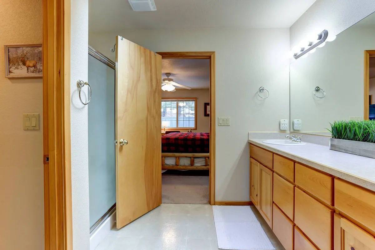 This image offers a view into a functional ensuite bathroom featuring wooden cabinets, a mirror, shower, and a glimpse of plaid bedding in the adjoining bedroom. The space is bright, comfortable, and conducive to relaxation and morning routines.