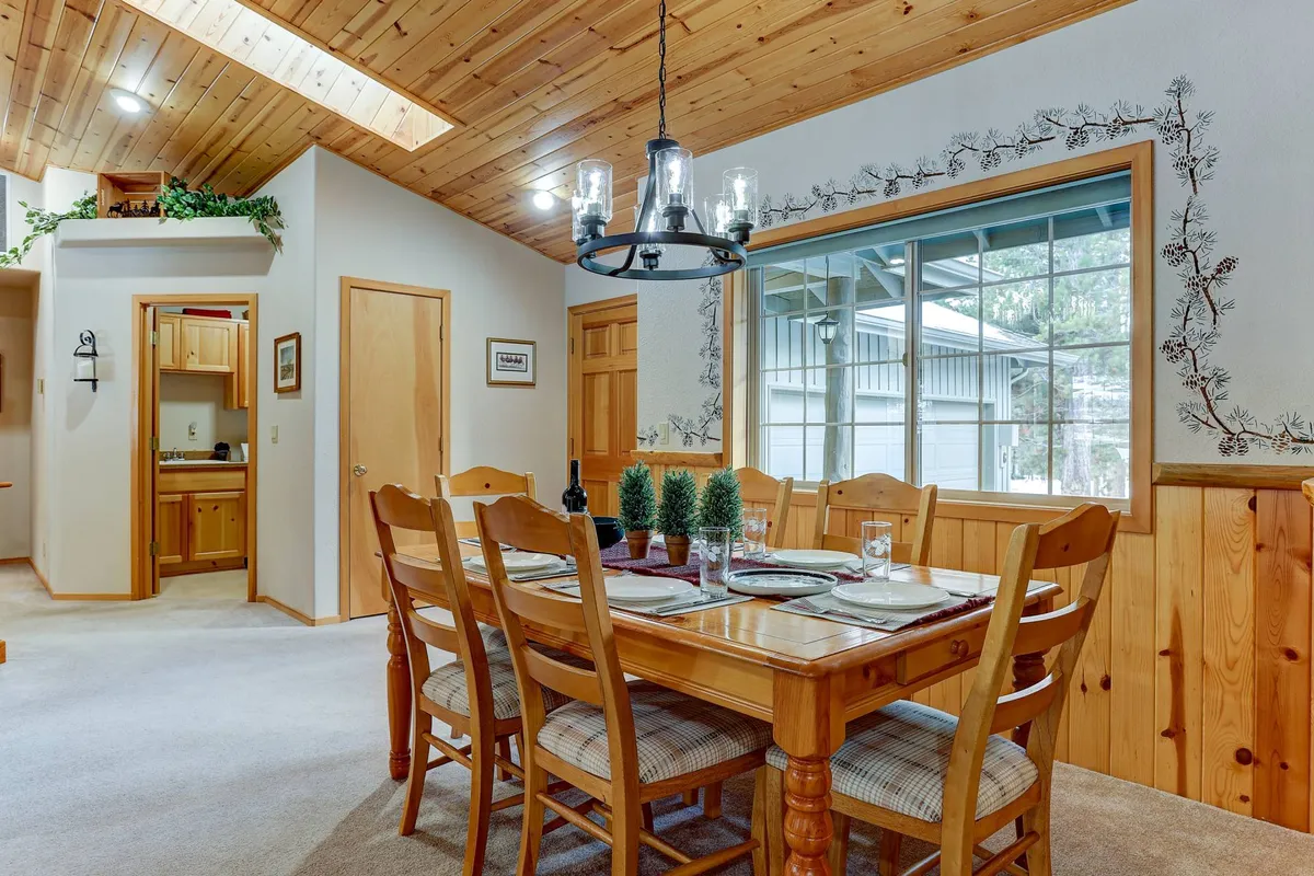 This image centers on the adjacent dining area, featuring a prominent dining table under a chandelier with a large window nearby. The scene is designed for group meals, spirited games, and celebrations, evoking a sense of togetherness and memorable moments.