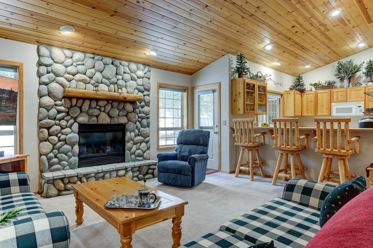 This image showcases a spacious living room with a prominent stone fireplace, soaring wooden ceilings, plaid sofas, and a view toward the kitchen bar. The inviting setup features cozy seating arranged for both relaxation and socializing, while natural light floods the space from large windows.