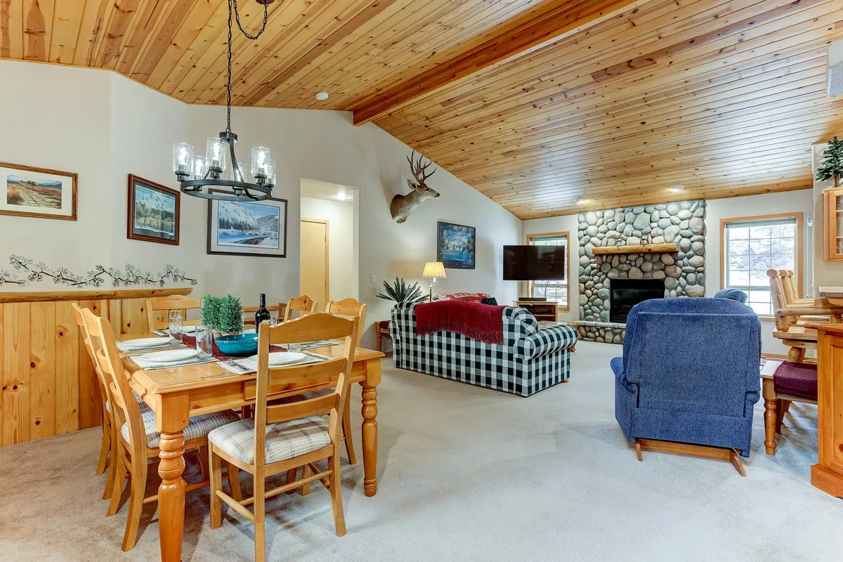 A wide perspective blends the dining area and living zone, highlighting the stone fireplace, wooden beams, and a plaid sofa. The scene radiates lodge charm and suggests easy transitions between meals, games, and fireside relaxation.