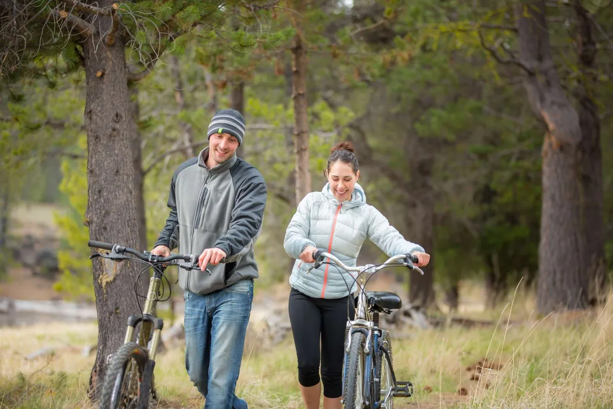 This exterior image features two bicycles, a couple walking together under tall trees, and a lush natural landscape. The mood is adventurous and joyful, capturing the essence of exploring scenic paths and spending active days outdoors.