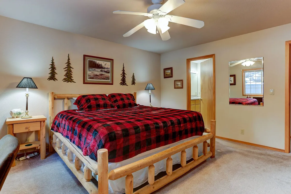 A comfortable bedroom is depicted here with a log bed frame, plaid bedspread, wall art, bedside lamps, and a mirrored wall. The atmosphere is warm and inviting, perfect for unwinding with a book or enjoying the morning sun.