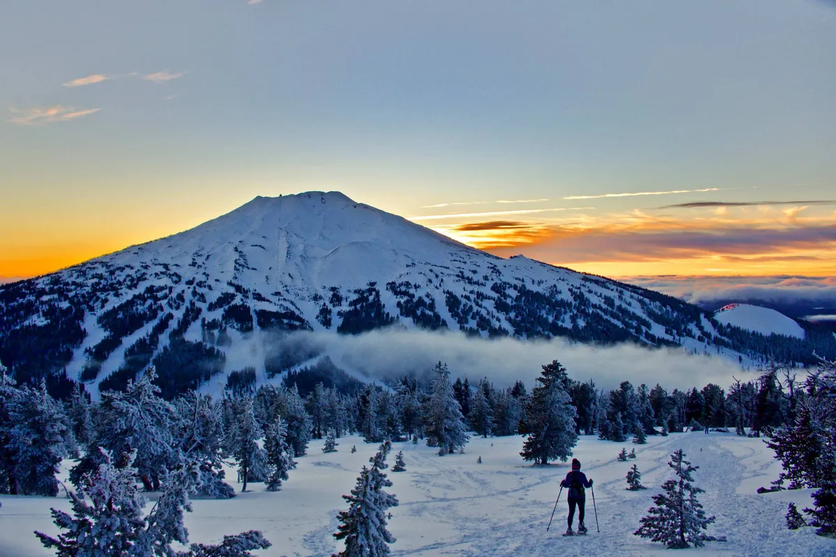 A breathtaking sunset view of a snow-covered mountain, with a skier in action and evergreen trees in the foreground. The scene is serene and invigorating, capturing the thrill of winter sports in a stunning setting.